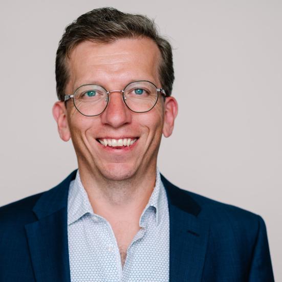 Kristian Steen Frederiksen, overlæge ved Hukommelsesklinikken på Nationalt Videnscenter for Demens, sidder i en lyseblåskjorte og en mørk blazer og smiler stort. Han er iført runde briller og sidder foran en neutral, beige baggrund. 