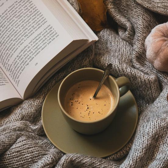 en kop kaffe og en åben bog ligger på et gråt strikket tæppe. I siden af billedet ses et lille efterårs græskar og et vissen blad