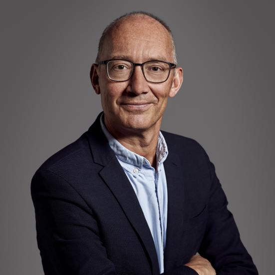 Et portræt af Niels Krause-Kjær. Han er iført en mørkeblå blazer og lyseblå skjorte og store sorte briller. Han står med korslagte arme og vender mod beskueren med et lille skævt smil på læben.