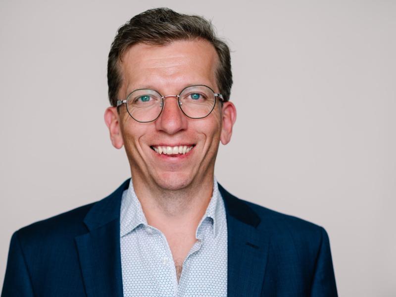 Kristian Steen Frederiksen, overlæge ved Hukommelsesklinikken på Nationalt Videnscenter for Demens, sidder i en lyseblåskjorte og en mørk blazer og smiler stort. Han er iført runde briller og sidder foran en neutral, beige baggrund.