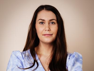 Portræt af Pernille Oldgaard, der står med armene over kors i en lyseblå feminin skjortebluse og langt, mørkt løst hår. Hun har et lille smil i mundvigen.