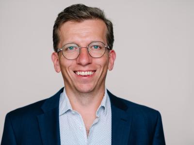 Kristian Steen Frederiksen, overlæge ved Hukommelsesklinikken på Nationalt Videnscenter for Demens, sidder i en lyseblåskjorte og en mørk blazer og smiler stort. Han er iført runde briller og sidder foran en neutral, beige baggrund. 