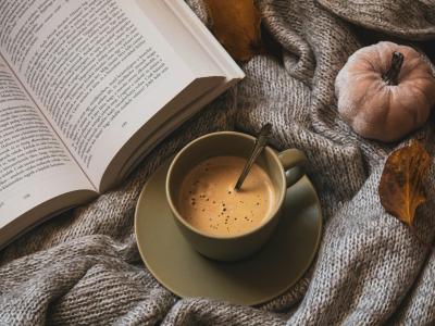 en kop kaffe og en åben bog ligger på et gråt strikket tæppe. I siden af billedet ses et lille efterårs græskar og et vissen blad
