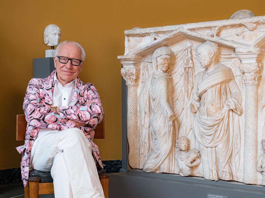 Ditlev Tamm in a pink floral blazer and white pants smiling while sitting in front of a plastor or marble.  relief