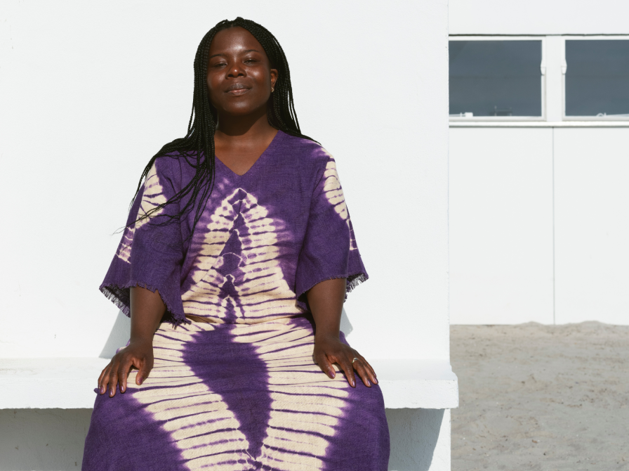 Lucia Odoom er en brun kvinde iført en lilla og beige batikfarvet løs kjole og sorte sandaler med hvide strømper. Hun smiler let til kameraet, mens hun sidder på en bænk foran en hvid væg i strålende solskin. I baggrunden anes et glimt af blå himmel og trætoppe.