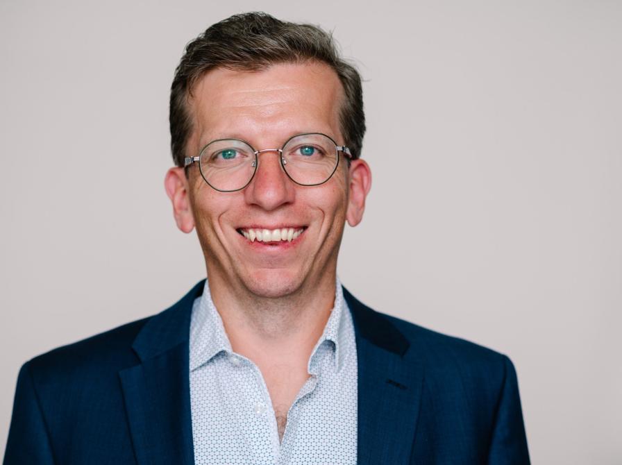 Kristian Steen Frederiksen, overlæge ved Hukommelsesklinikken på Nationalt Videnscenter for Demens, sidder i en lyseblåskjorte og en mørk blazer og smiler stort. Han er iført runde briller og sidder foran en neutral, beige baggrund.